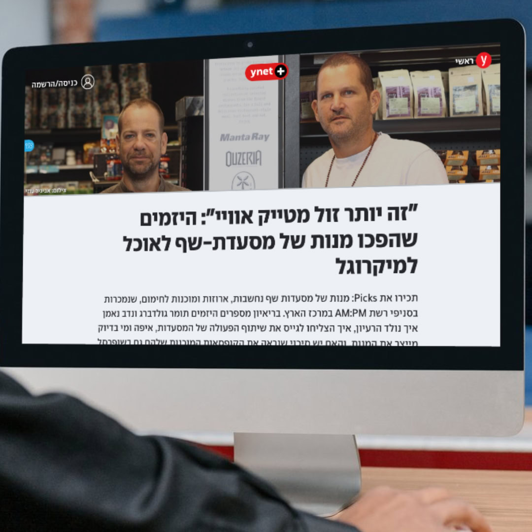 אדם מסתכל על מסך מחשב המציג מאמר מקוון מתוך "ynet". הכתבה מציגה שני גברים עומדים מול מדפים. הכותרת, בעברית, דנה כיצד יזמים אלה הפכו את ארוחות המסעדות למוצרי מזון בסופרמרקט, ויצרו עסק פורח.